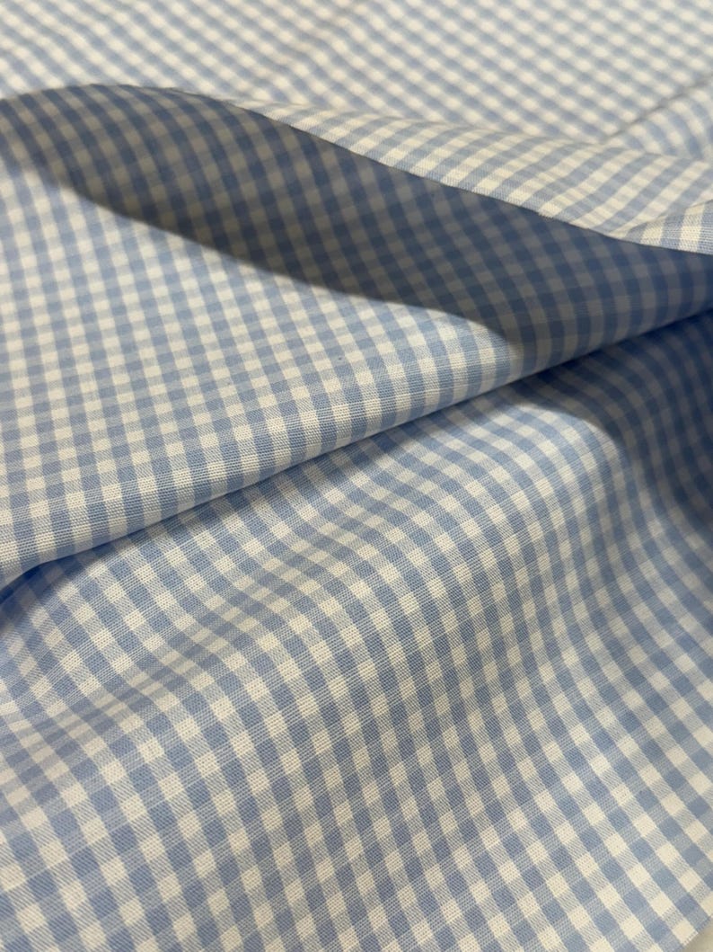 May include: Close-up of a light blue and white gingham fabric. The fabric is folded, showing the texture and pattern of the woven material. The pattern is a classic checkered design, with small squares of light blue and white.