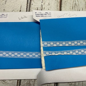 May include: Two fabric swatches with white lace trim. The swatch on the left is a bright blue color and the swatch on the right is a light blue color. The text on the swatches reads "N° 7.78 Mts. 33. Coton 90% Yds. 76.6 Nylon 10% Made in France" and "N° 7.78 Mts. 35 Coton 90% Yds. 8.0 Nylon 10% Made in France". The text "White" is written on the left swatch and "Champagne Ivory" is written on the right swatch.