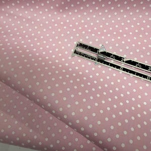 May include: A light pink fabric featuring a repeating pattern of small white polka dots. A metal measuring tool is placed on the fabric, showing a measurement of approximately 3 inches across the visible scale of the tool.