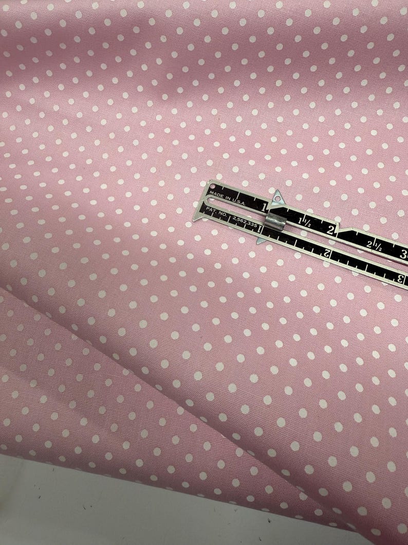 May include: A light pink fabric featuring a repeating pattern of small white polka dots. A metal measuring tool is placed on the fabric, showing a measurement of approximately 3 inches across the visible scale of the tool.