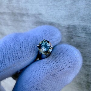 May include: A round, blue gemstone with a silver setting. The gemstone has a slight green hue and is surrounded by four prongs.
