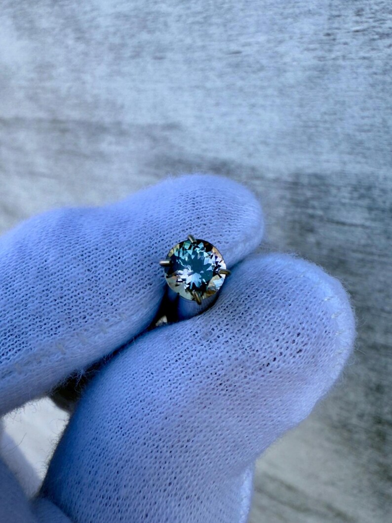 May include: A round, blue gemstone with a silver setting. The gemstone has a slight green hue and is surrounded by four prongs.