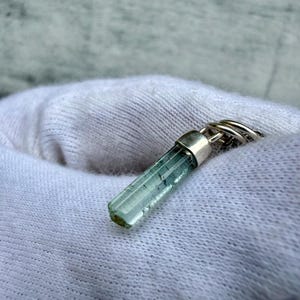 May include: A light blue tourmaline crystal pendant with a silver cap and ring. The crystal is cylindrical with visible internal structures. The pendant is displayed on a white fabric background.
