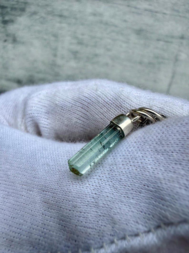 May include: A light blue tourmaline crystal pendant with a silver cap and ring. The crystal is cylindrical with visible internal structures. The pendant is displayed on a white fabric background.