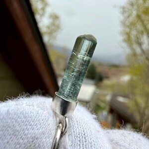 May include: A blue-green tourmaline crystal pendant with a silver setting. The crystal is cylindrical with visible internal striations. The pendant is attached to a silver chain. The background is blurred, showing a natural outdoor setting.
