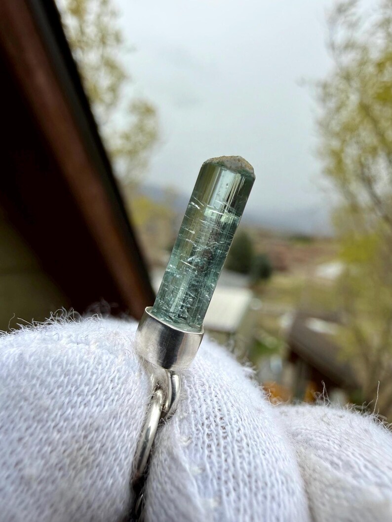 May include: A blue-green tourmaline crystal pendant with a silver setting. The crystal is cylindrical with visible internal striations. The pendant is attached to a silver chain. The background is blurred, showing a natural outdoor setting.