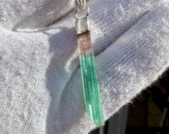 May include: A pendant necklace with a bi-colour tourmaline gemstone. The gemstone transitions from pinkish-brown to green. The pendant hangs from a silver-toned chain. The necklace is displayed against a white background.