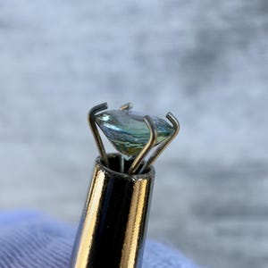 May include: A light blue gemstone held in a metal prong setting. The gemstone is being held by a metal tool with a silver finish.