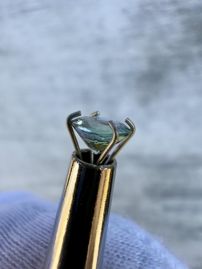 May include: A light blue gemstone held in a metal prong setting. The gemstone is being held by a metal tool with a silver finish.