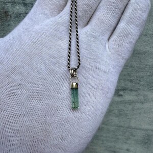 May include: A silver necklace featuring a small, light green, cylindrical gemstone pendant. The pendant hangs from a delicate, twisted silver chain. The gemstone is set in silver.