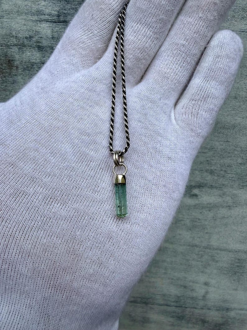 May include: A silver necklace featuring a small, light green, cylindrical gemstone pendant. The pendant hangs from a delicate, twisted silver chain. The gemstone is set in silver.