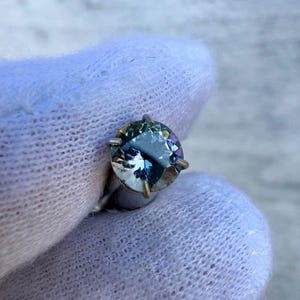 May include: A round, faceted gemstone with a blue-green hue. The gemstone is set in a silver prong setting.