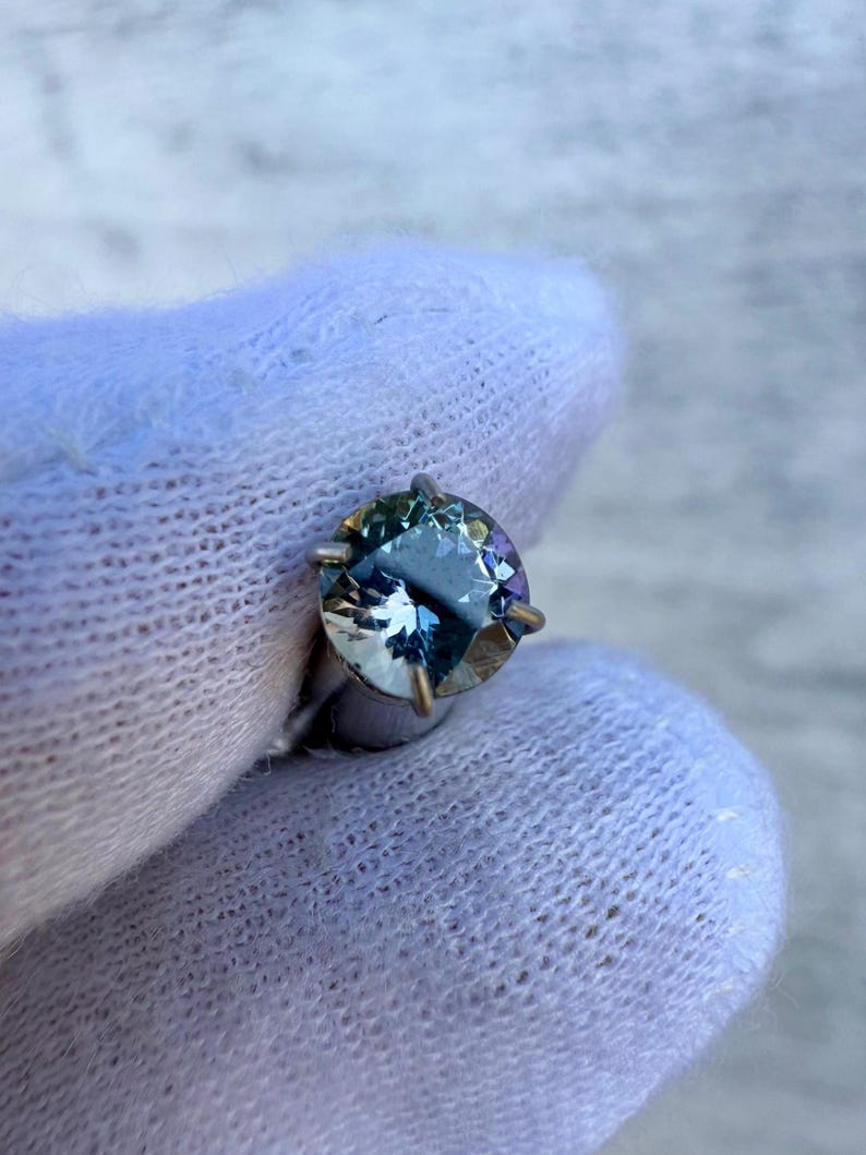 May include: A round, faceted gemstone with a blue-green hue. The gemstone is set in a silver prong setting.