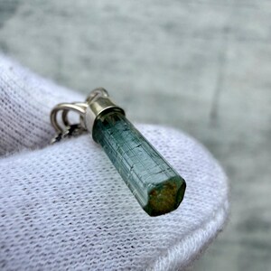 May include: A pendant featuring a raw, hexagonal, blue-green tourmaline crystal set in a silver-toned metal cap. The elongated, translucent gemstone displays natural vertical striations along its length and a flat, textured base.