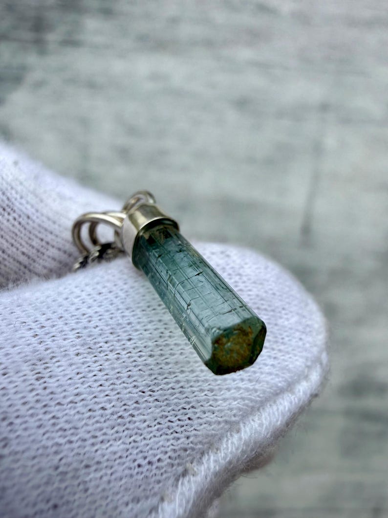 May include: A pendant featuring a raw, hexagonal, blue-green tourmaline crystal set in a silver-toned metal cap. The elongated, translucent gemstone displays natural vertical striations along its length and a flat, textured base.