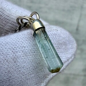 May include: A pendant featuring a long, slender, pale blue-green crystal point encased in a polished silver cap. The crystal has a hexagonal shape with visible internal inclusions and a small brown tip at the base, suspended from a silver chain.