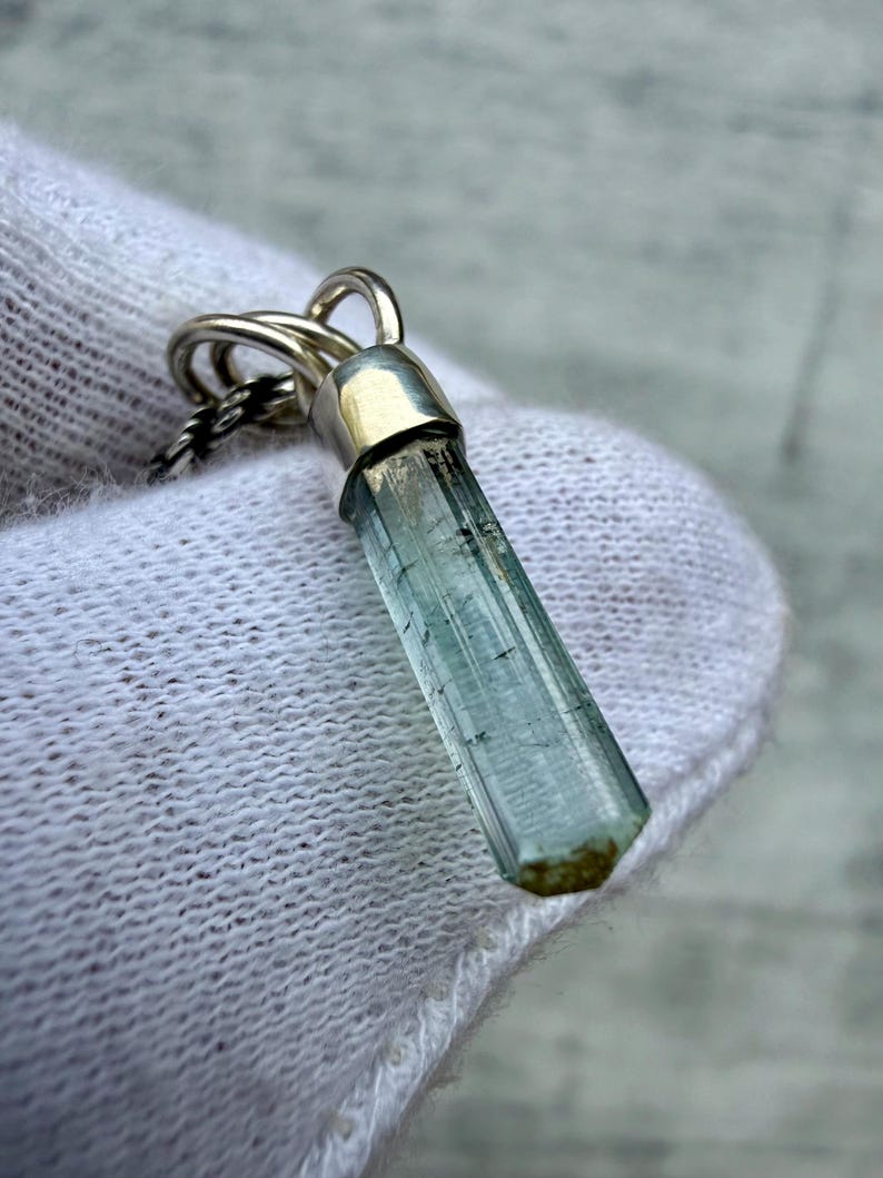 May include: A pendant featuring a long, slender, pale blue-green crystal point encased in a polished silver cap. The crystal has a hexagonal shape with visible internal inclusions and a small brown tip at the base, suspended from a silver chain.