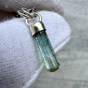 May include: A pendant featuring a raw, elongated, translucent blue-green tourmaline crystal held by a polished silver cap. The crystal has visible vertical striations and is attached to a silver jump ring connected to a thin, dark silver chain.