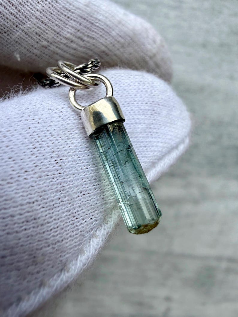 May include: A pendant featuring a raw, elongated, translucent blue-green tourmaline crystal held by a polished silver cap. The crystal has visible vertical striations and is attached to a silver jump ring connected to a thin, dark silver chain.