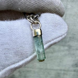 May include: A silver necklace with a pendant featuring a light green, cylindrical gemstone. The gemstone is encased in a silver cap and hangs from a silver chain. The pendant has a natural, textured appearance.