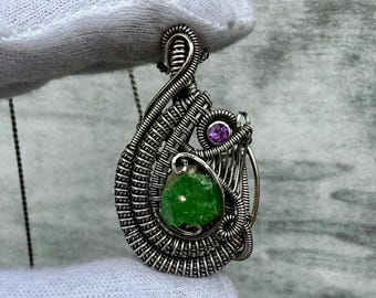 May include: A silver wire wrapped pendant featuring a green gemstone and a smaller purple gemstone. The pendant is hanging from a silver chain.