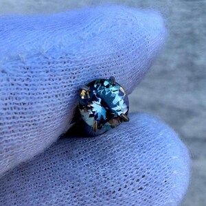 May include: A round, faceted gemstone with a blue-green hue. The gemstone is set in a silver-toned setting with four prongs.