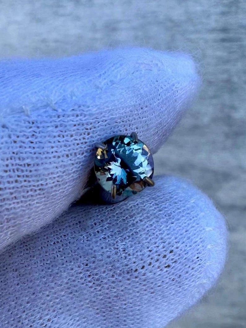 May include: A round, faceted gemstone with a blue-green hue. The gemstone is set in a silver-toned setting with four prongs.