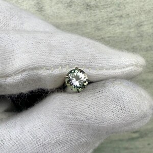 May include: A round, clear gemstone with a green hue, set in a silver prong setting. The gemstone is held in a white gloved hand.