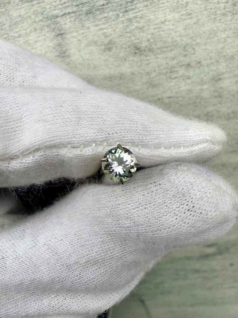 May include: A round, clear gemstone with a green hue, set in a silver prong setting. The gemstone is held in a white gloved hand.