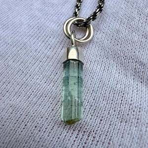 May include: A silver necklace with a pendant featuring a pale blue, cylindrical gemstone. The gemstone is set in a silver cap and suspended from a silver chain. The background is a white, textured fabric.