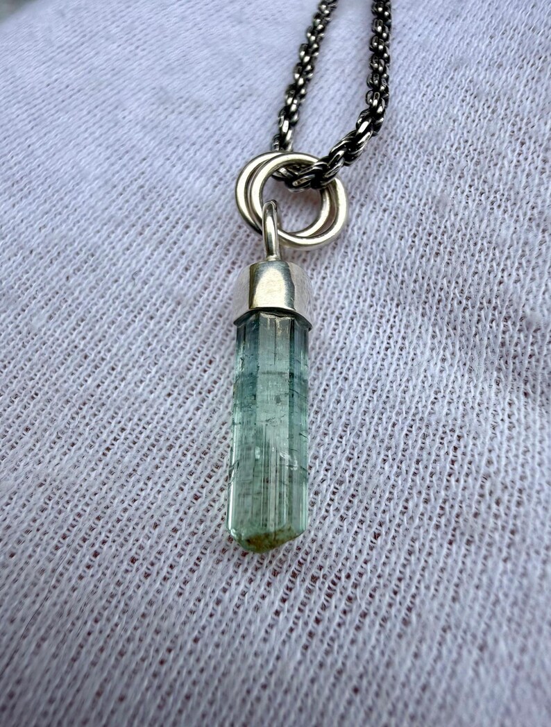 May include: A silver necklace with a pendant featuring a pale blue, cylindrical gemstone. The gemstone is set in a silver cap and suspended from a silver chain. The background is a white, textured fabric.