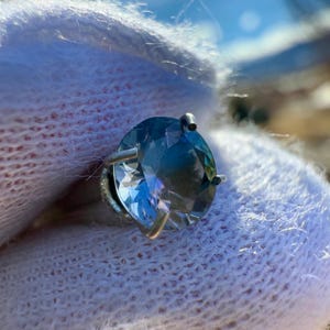 May include: A round, faceted, blue gemstone set in a silver ring setting. The ring is on a white fluffy surface.