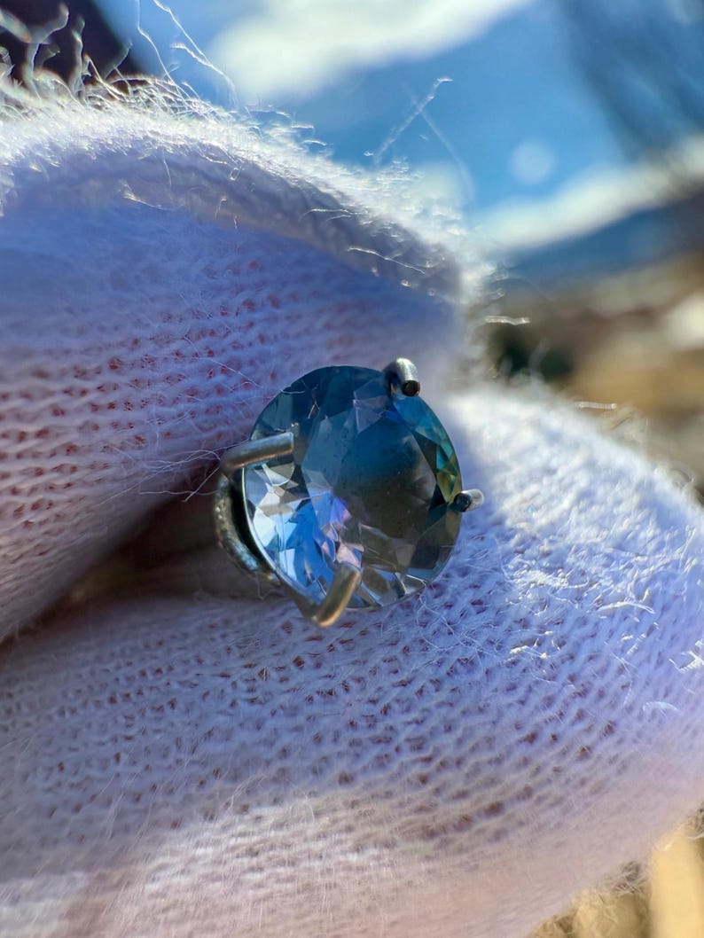 May include: A round, faceted, blue gemstone set in a silver ring setting. The ring is on a white fluffy surface.