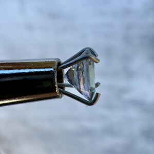 May include: A clear gemstone held in a metal prong setting. The gemstone is faceted and has a slight blue hue.