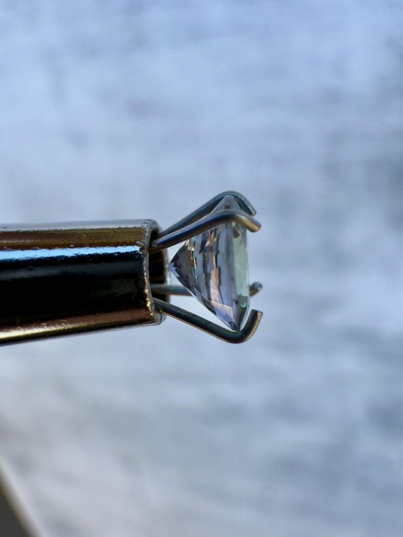 May include: A clear gemstone held in a metal prong setting. The gemstone is faceted and has a slight blue hue.