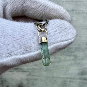 May include: A pendant featuring a long, translucent, light green tourmaline crystal encased in a polished silver cap. The crystal hangs from a silver jump ring attached to a dark, braided metal chain, showcasing a vertical, faceted structure.