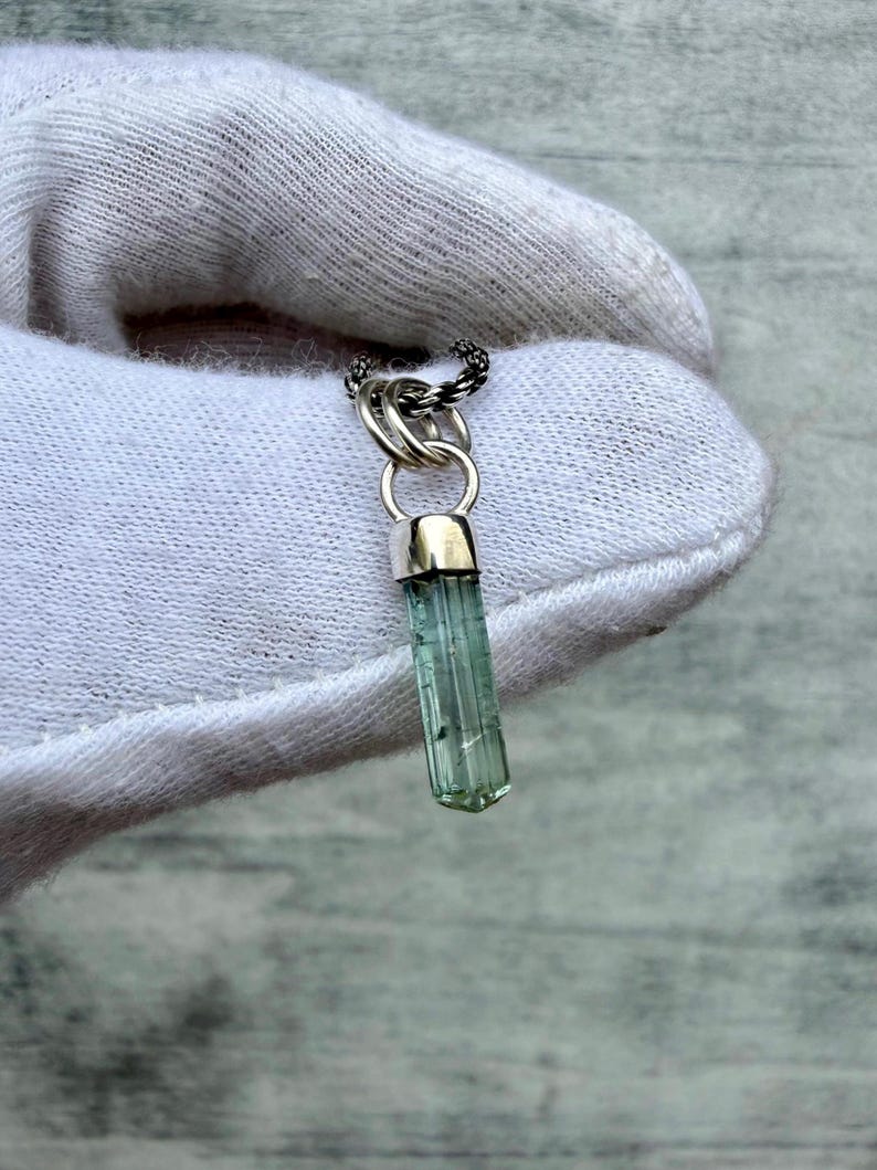 May include: A pendant featuring a long, translucent, light green tourmaline crystal encased in a polished silver cap. The crystal hangs from a silver jump ring attached to a dark, braided metal chain, showcasing a vertical, faceted structure.