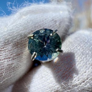May include: A round, faceted, green gemstone ring with a silver setting. The gemstone has a blue-green hue and a slight purple tint.