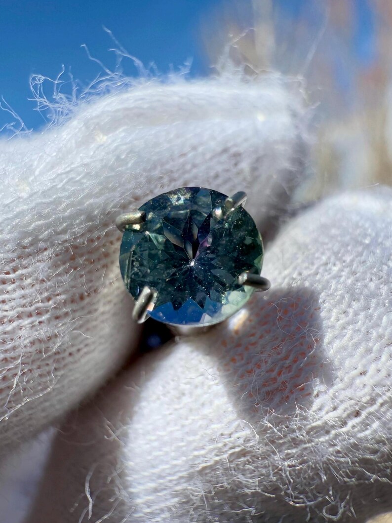 May include: A round, faceted, green gemstone ring with a silver setting. The gemstone has a blue-green hue and a slight purple tint.