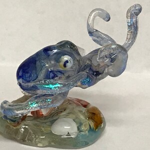 May include: A blue glass sculpture of an octopus with iridescent details, perched on a clear glass base with colorful swirls and a white shell.