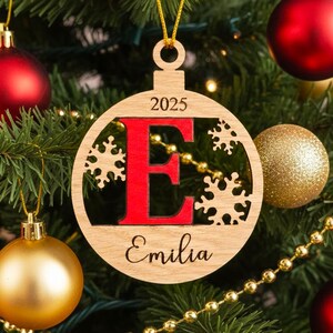 May include: Wooden Christmas bauble with a red letter "E" and the name "Emilia" below. The year "2025" is above the letter. The bauble hangs on a Christmas tree with red and gold decorations.