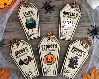 May include: Wooden coffin-shaped Halloween basket tags with names and spooky illustrations. Each tag features a name, the words "BOO BASKET", and a different Halloween-themed graphic, such as a frog, ghost, bat, pumpkin, and cat.