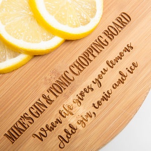Puede incluir: Una tabla de cortar redonda de madera con el texto "MIKE'S GIN & TONIC CHOPPING BOARD" y "When life gives you lemons Add gin, tonic and ice" grabado en ella. Hay cuatro rodajas de lim&oacute;n en la tabla.