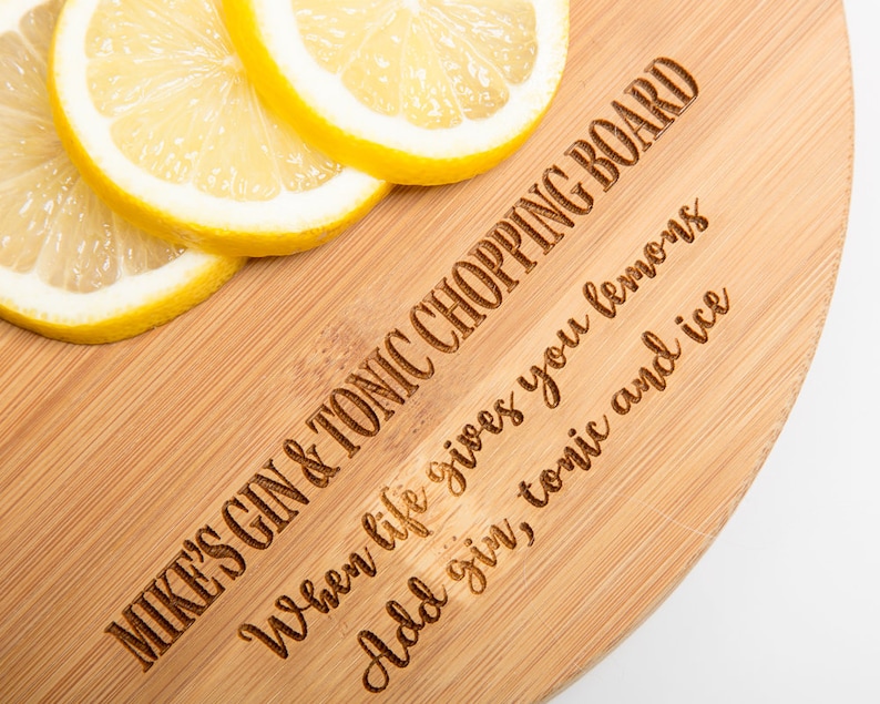 Puede incluir: Una tabla de cortar redonda de madera con el texto "MIKE'S GIN & TONIC CHOPPING BOARD" y "When life gives you lemons Add gin, tonic and ice" grabado en ella. Hay cuatro rodajas de lim&oacute;n en la tabla.