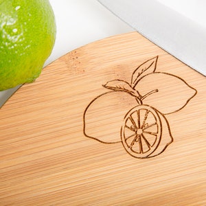 Puede incluir: Una tabla de cortar redonda de bamb&uacute; con un dise&ntilde;o de limones grabado con l&aacute;ser. La tabla es de color marr&oacute;n claro y tiene una veta de madera natural.