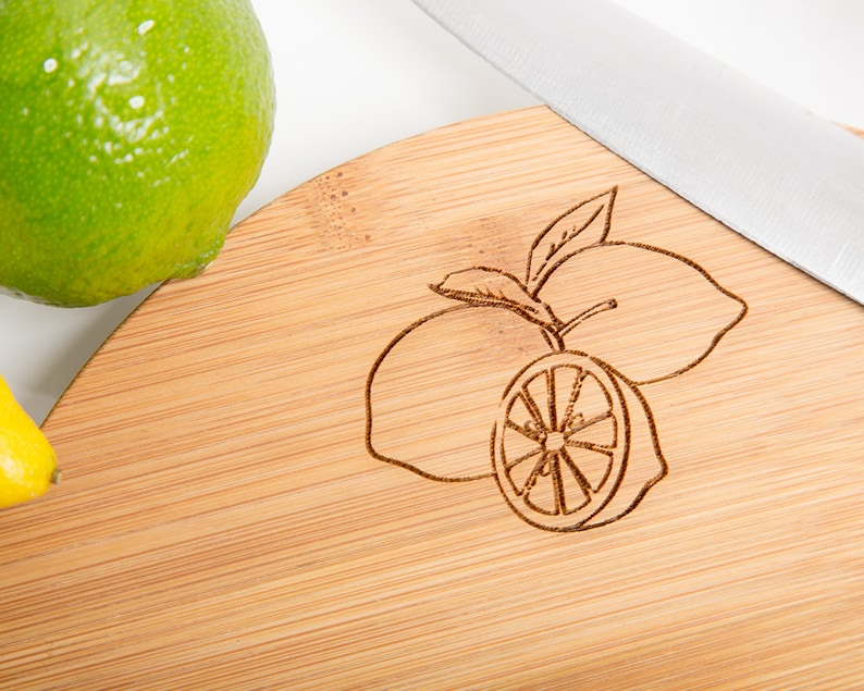 Puede incluir: Una tabla de cortar redonda de bamb&uacute; con un dise&ntilde;o de limones grabado con l&aacute;ser. La tabla es de color marr&oacute;n claro y tiene una veta de madera natural.