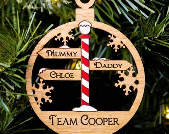 May include: Wooden Christmas ornament with a red ribbon. The ornament features a signpost design with the words "Mummy," "Chloe," and "Daddy" on directional arrows. The words "Team Cooper" are at the bottom.