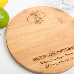Puede incluir: Una tabla de cortar redonda de madera con un acabado marr&oacute;n claro. La tabla est&aacute; grabada con el texto "MIKE'S GIN & TONIC CHOPPING BOARD" y una peque&ntilde;a imagen de limones. El texto debajo del nombre de la tabla dice "When life gives you lemons Add gin, tonic and ice."