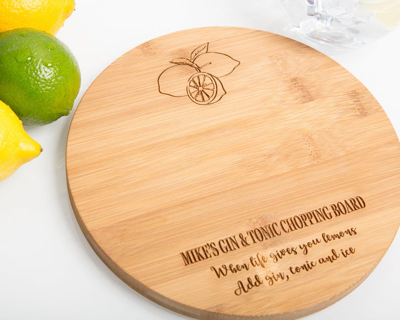 Puede incluir: Una tabla de cortar redonda de madera con un acabado marr&oacute;n claro. La tabla est&aacute; grabada con el texto "MIKE'S GIN & TONIC CHOPPING BOARD" y una peque&ntilde;a imagen de limones. El texto debajo del nombre de la tabla dice "When life gives you lemons Add gin, tonic and ice."