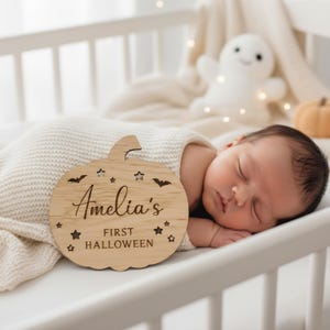 May include: A wooden pumpkin-shaped sign with the text "Amelia's First Halloween" and star and bat cutouts. The sign is placed next to a sleeping baby in a cot. A plush ghost and pumpkin are in the background.